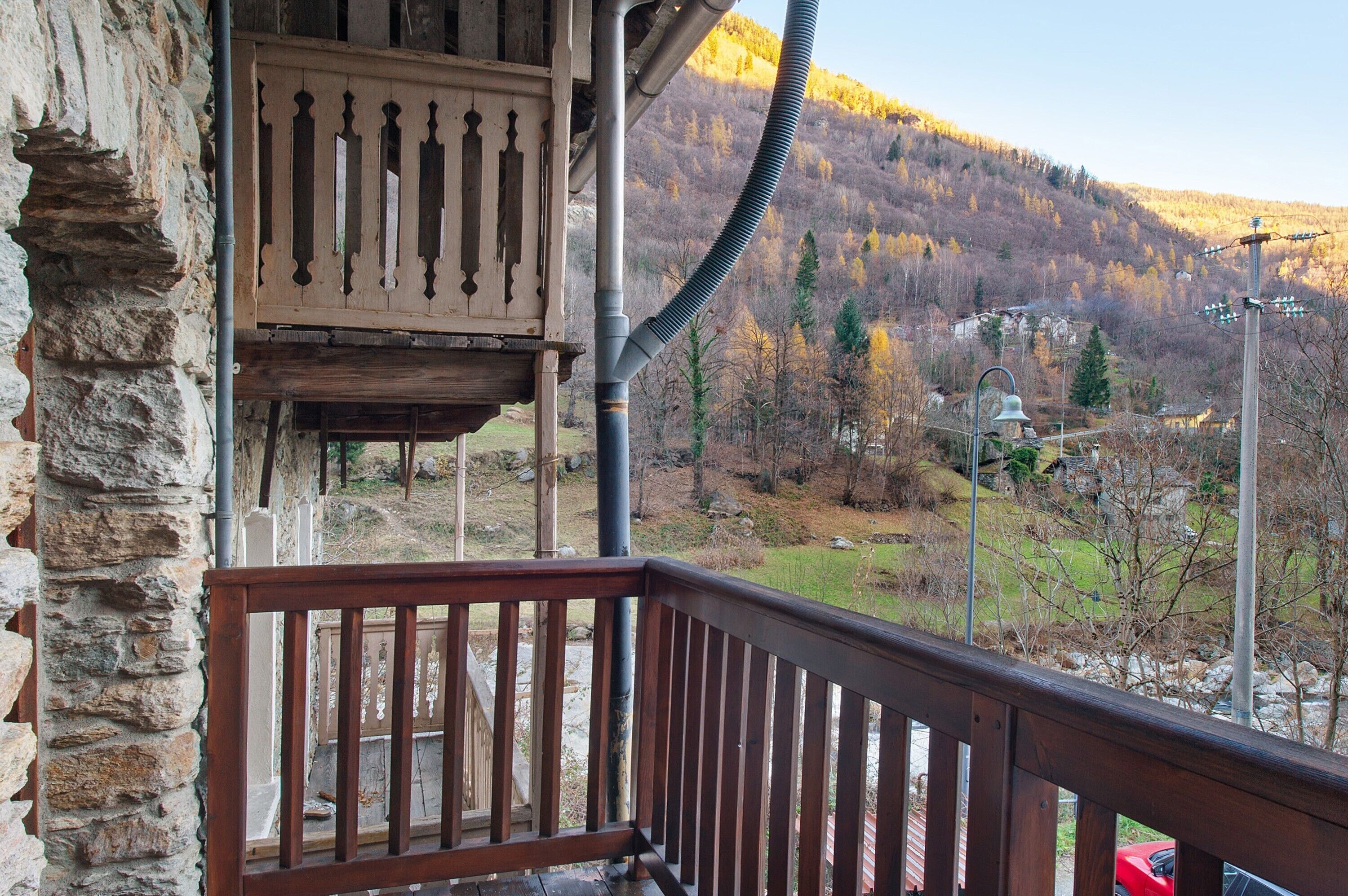 Ein Steinhaus mit einem Holzbalkon, Blick auf ein Tal mit Bäumen und ein kleines Gebäude, sowie ein rotes Auto vor der Fassade.