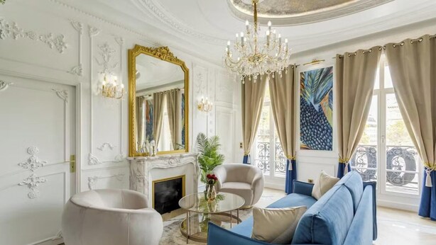 Een weelderige woonkamer met een witte open haard, een blauwe bank, een ronde salontafel en een grote spiegel met gouden lijst.