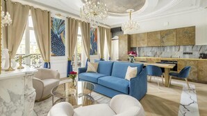 Een ruime woonkamer met een blauwe bank, witte fauteuils en een glazen salontafel. Achtergrond toont een keuken met een marmeren spatwand en moderne apparatuur.