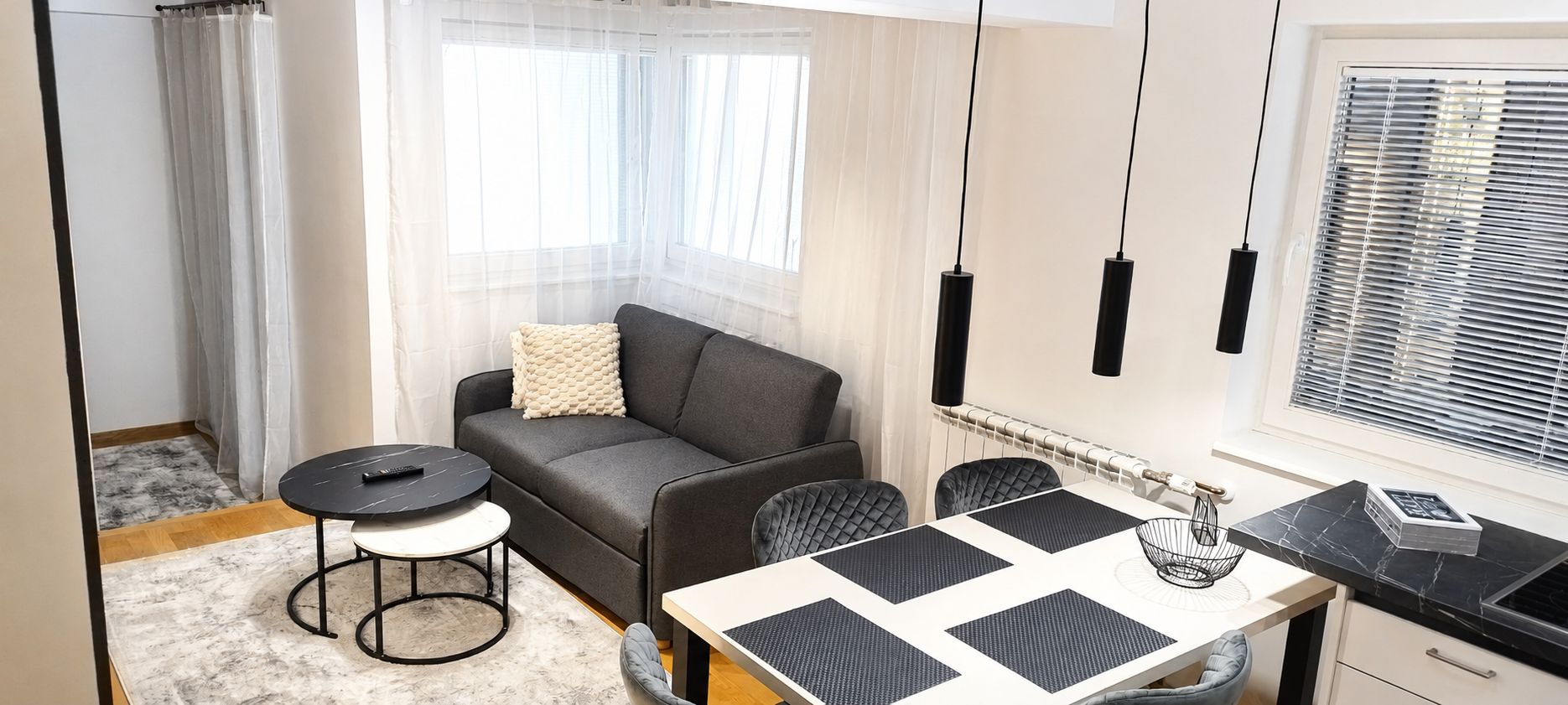 Een knus woongedeelte met een bank, een rond bijzettafeltje, een eettafel met stoelen en een keuken met fornuis.