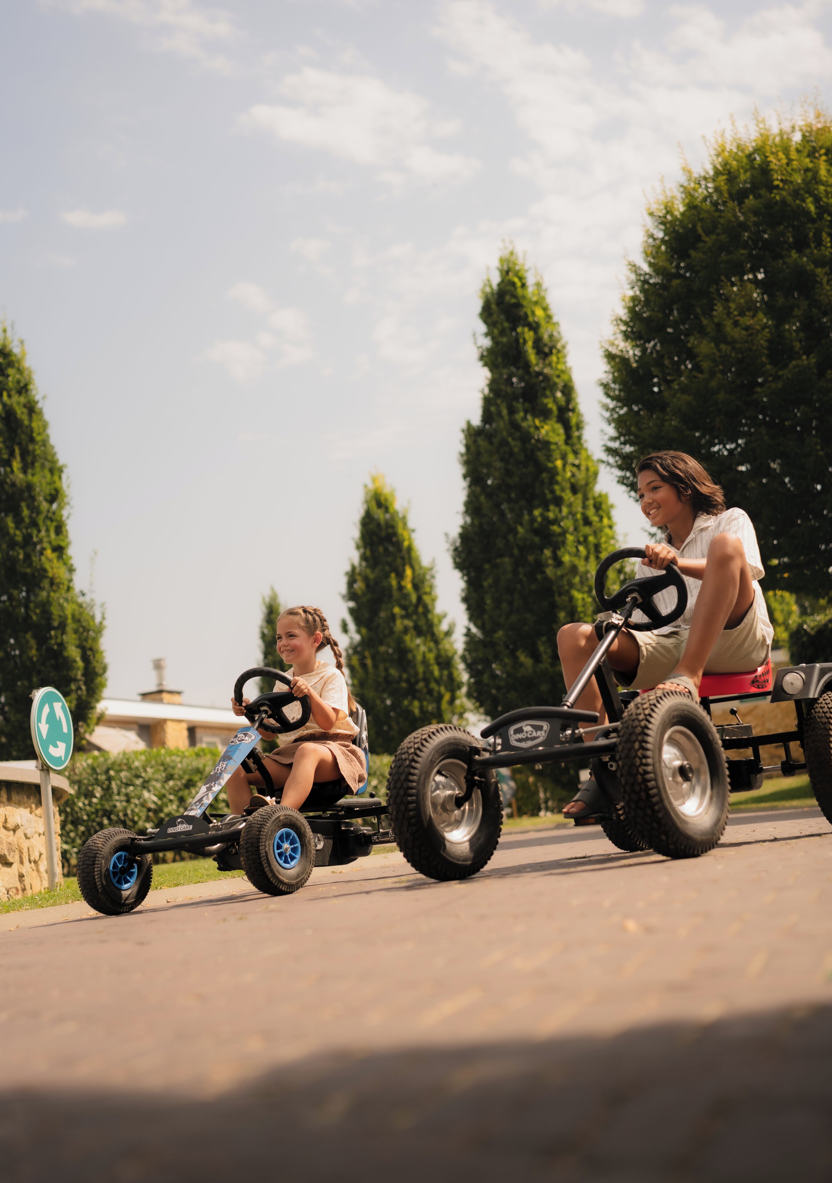 Two people on go-karts on a paved road with trees and a sign in the background.