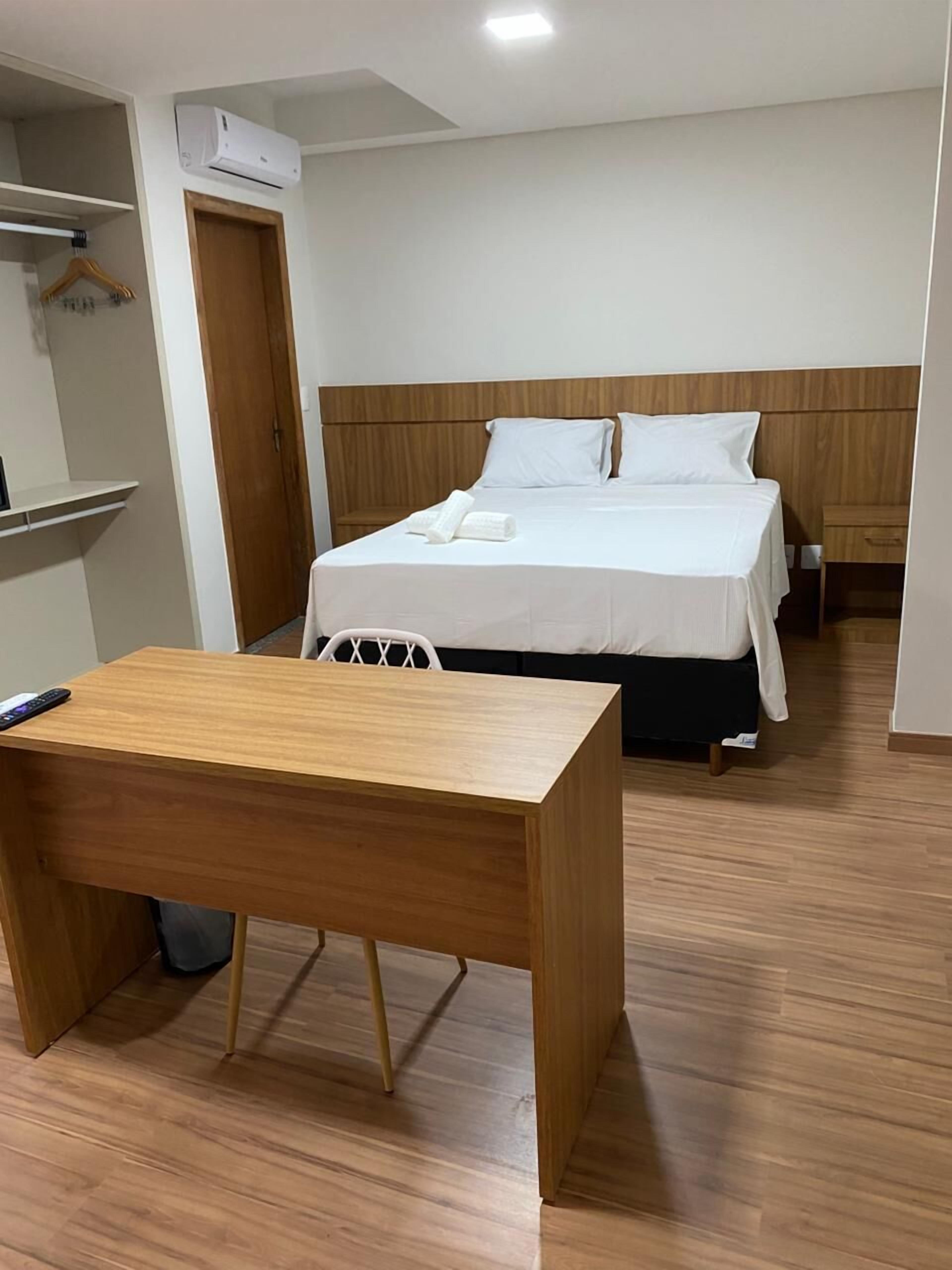 Habitación de hotel con un escritorio de madera, cama con ropa blanca, cabecera de madera y una silla blanca.