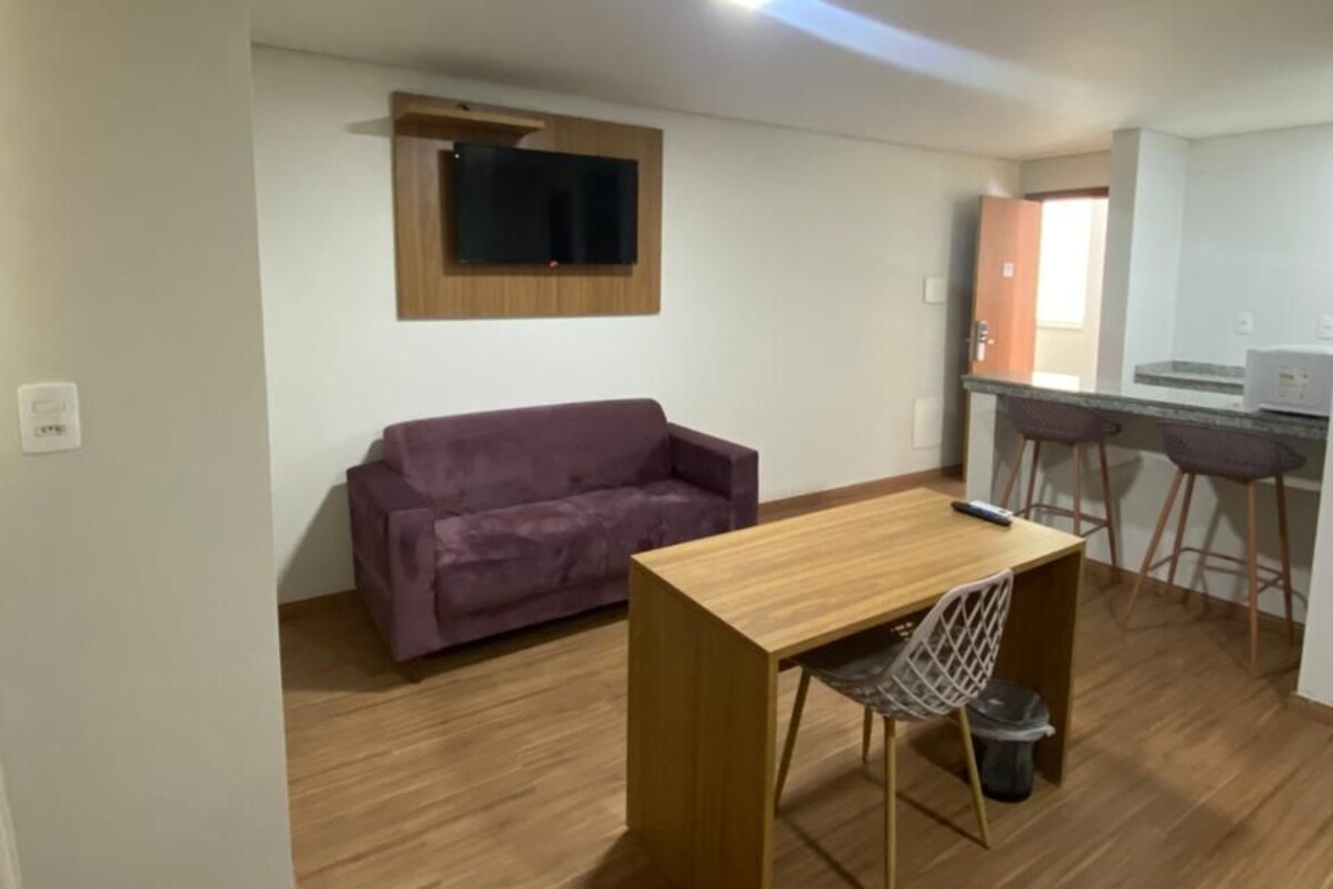 A compact living space with a wooden floor, a purple sofa, a small wooden table with a chair, a TV mounted on the wall, and a kitchenette area.