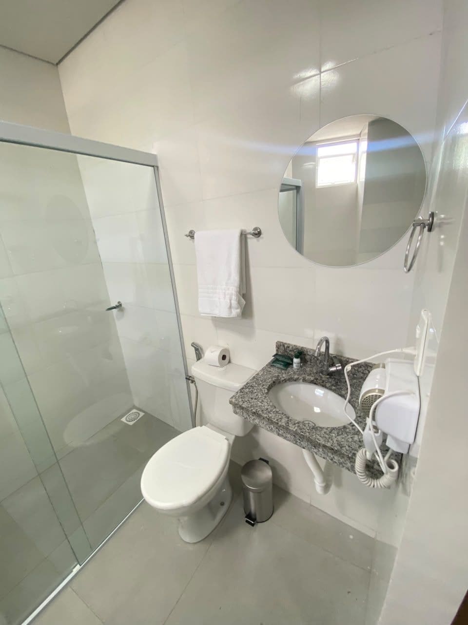 Un baño moderno con cabina de ducha de vidrio, un inodoro blanco, un lavamanos con encimera de granito, un espejo y un cubo de basura.