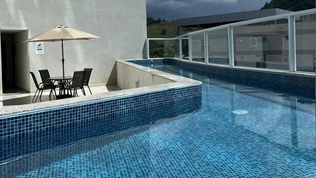 A swimming pool with tiled flooring, a table with chairs, and a glass-enclosed area. - San Diego Suites Manhuaçu (Manhuaçu)