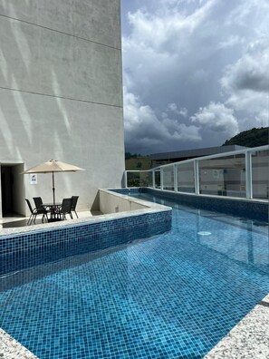 A swimming pool with tiled flooring, a table with chairs, and a glass-enclosed area. - San Diego Suites Manhuaçu (Manhuaçu)