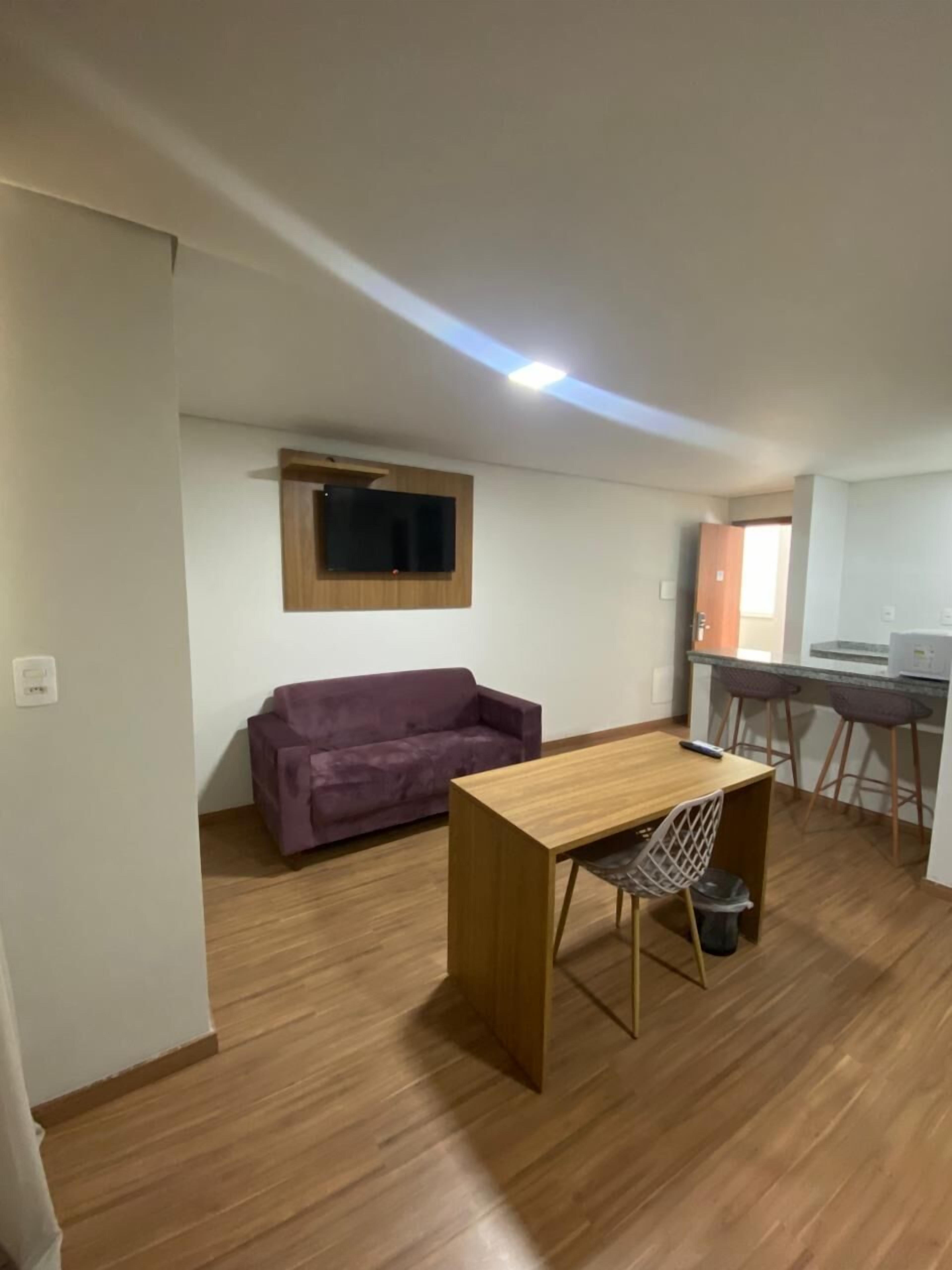Un espacio habitable compacto con piso de madera, un sofá morado, una pequeña mesa de madera con una silla, un televisor montado en la pared y una zona de kitchenette.