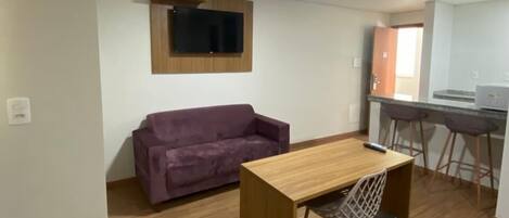 Un espacio habitable compacto con piso de madera, un sofá morado, una pequeña mesa de madera con una silla, un televisor montado en la pared y una zona de kitchenette.