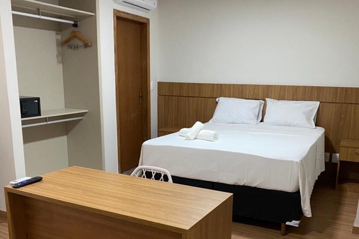 A hotel room with a wooden desk, a bed with white bedding, a wooden headboard, a wardrobe, and an air conditioning unit.