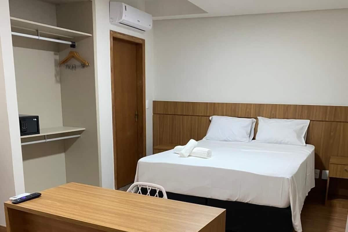 A hotel room with a wooden desk, a bed with white linens, a wooden headboard, and a wardrobe with a hanging rack.
