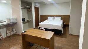 A hotel room with a wooden desk, a bed with white linens, and a small kitchenette area. - San Diego Suites Manhuaçu (Manhuaçu)