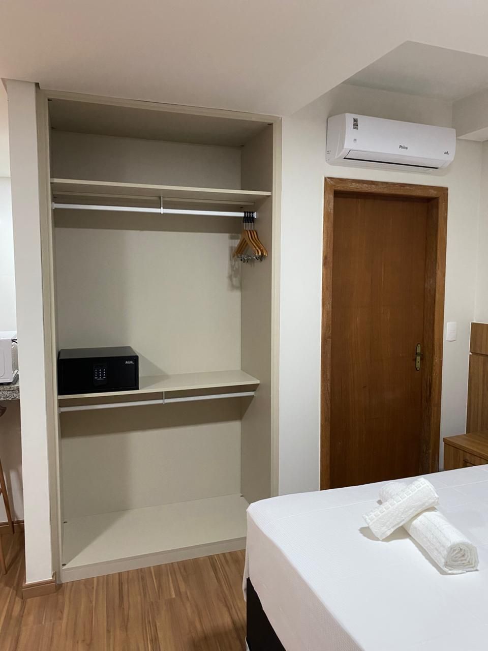 A bedroom with a bed, a wooden door, a white air conditioning unit, and a wardrobe with hanging clothes.