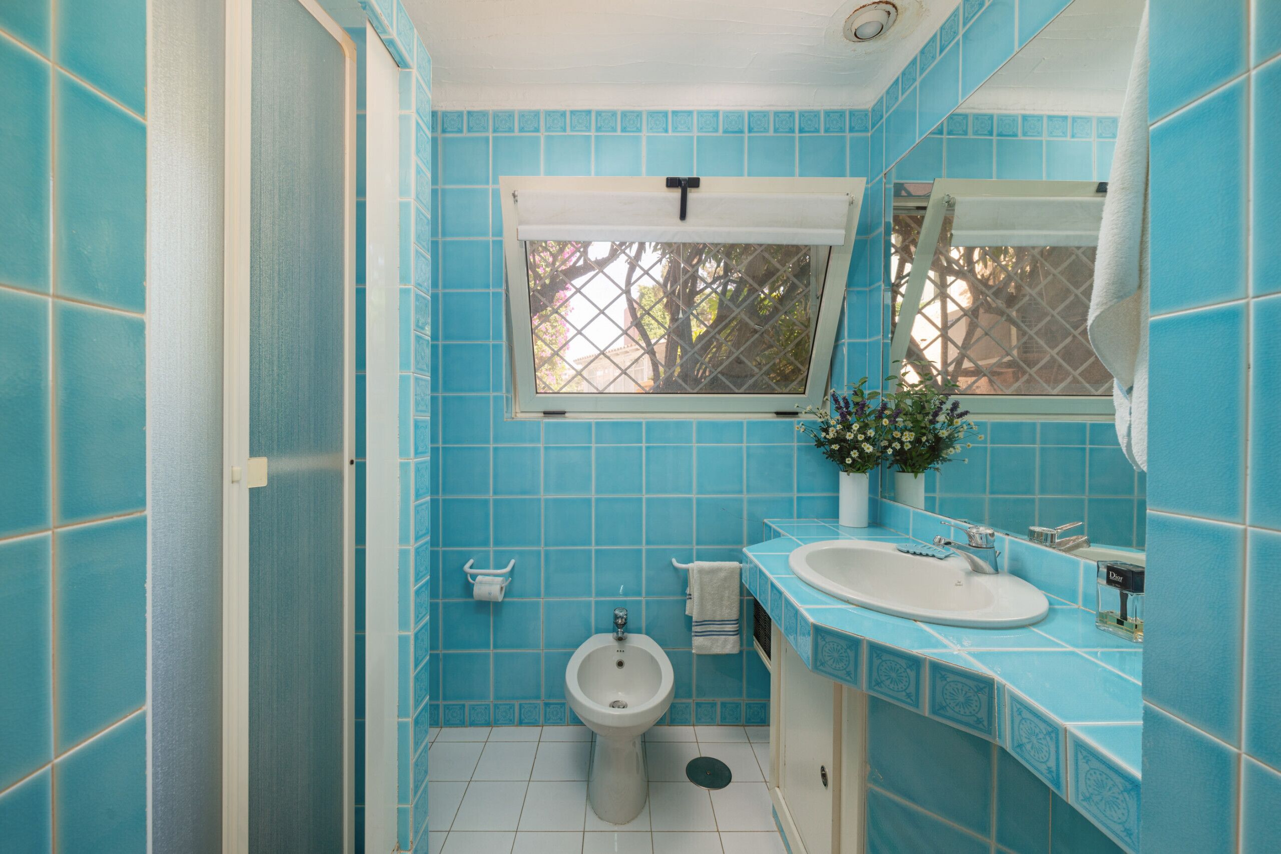 A bathroom with blue tiles, a white sink, a bidet, and a window with a view of trees.