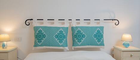 Una cama bien hecha con sábanas blancas, dos almohadones decorativos a juego, una lámpara de noche y una mesita de noche con lámpara.