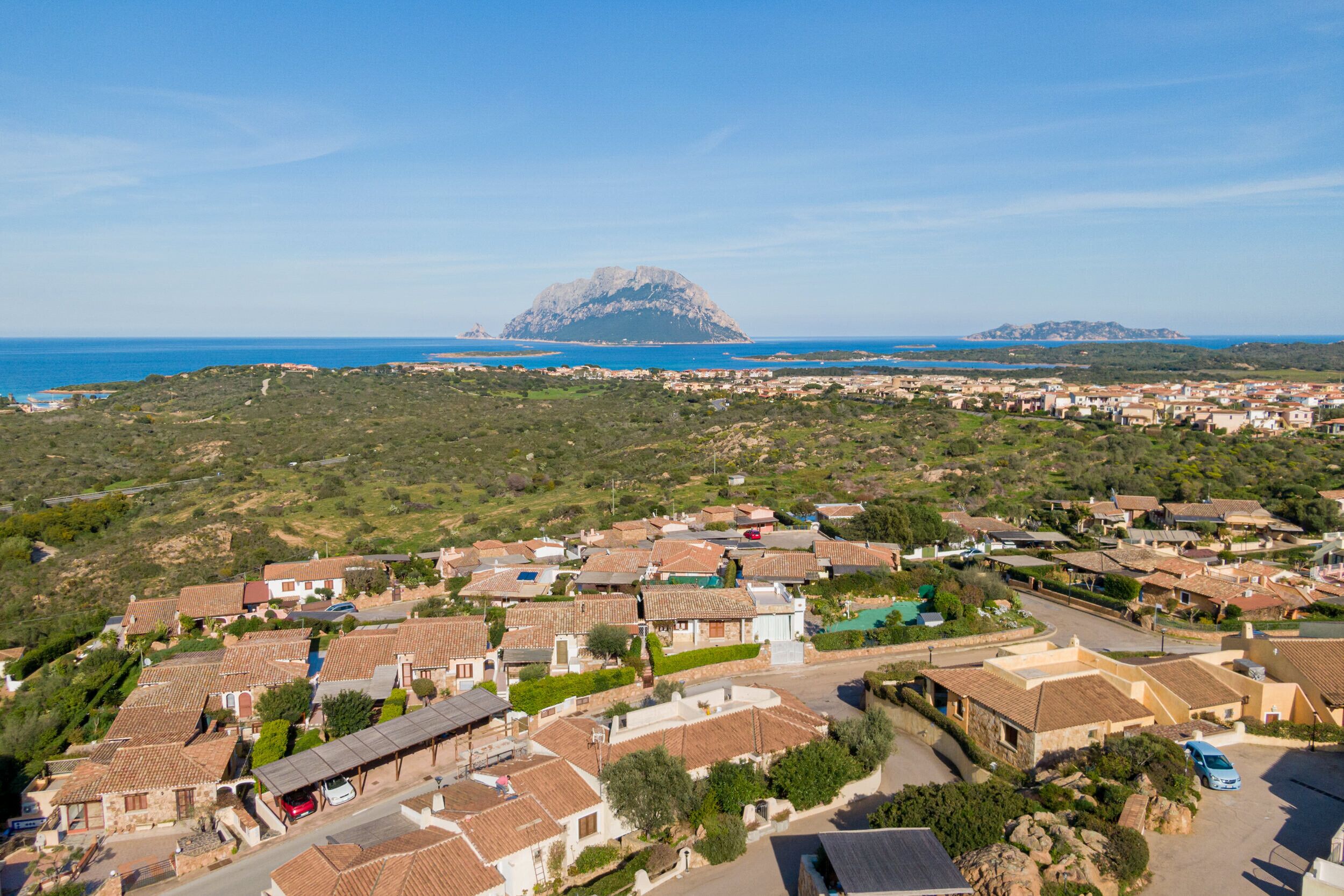 A coastal town with terracotta-roofed houses, a prominent mountain in the background, and a clear blue sky.
