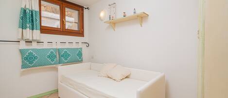 A small, white room with a single white sofa, a window with curtains, a wall-mounted shelf with decorative items, and a door.