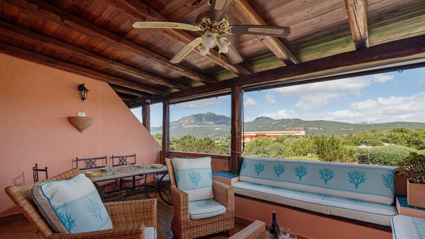 A covered patio with wicker furniture, a table, and a view of mountains. - Casa Graziana by Wonderful Italy (Olbia)