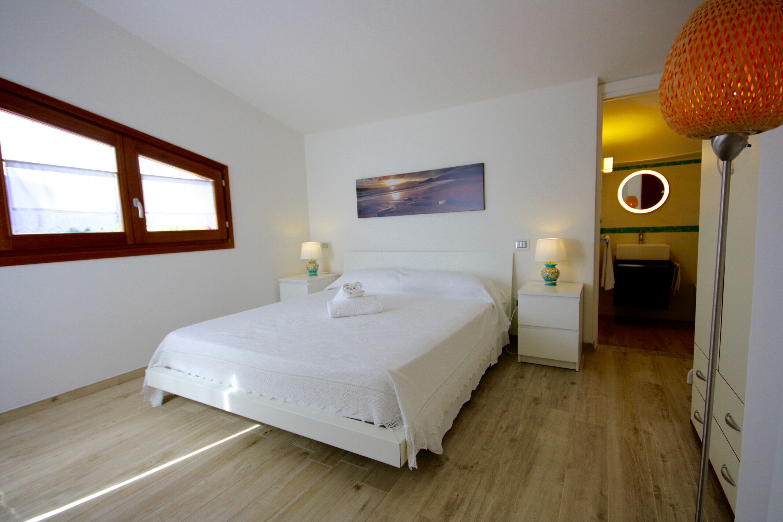 Une chambre à coucher avec un lit, des tables de chevet, une fenêtre, un tableau, et une salle de bain visible à travers une porte ouverte.