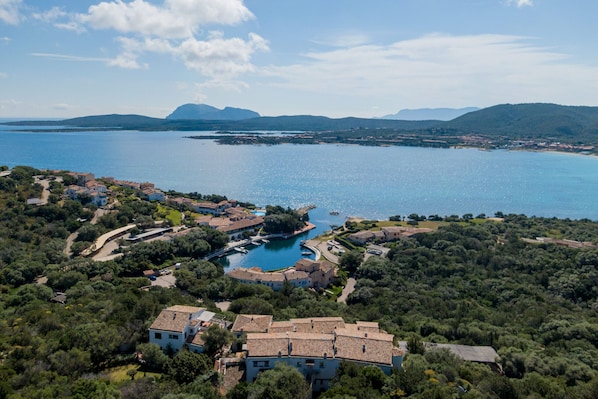 An aerial view of a coastal property with a swimming pool surrounded by buildings, a marina, and a backdrop of mountains and the sea. - Casa Pierina by Wonderful Italy (Olbia)