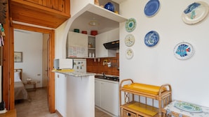 Una cucina compatta con pensili bianchi, un piano di lavoro in legno e un piccolo angolo ristoro con tavolo e sedie in vimini.