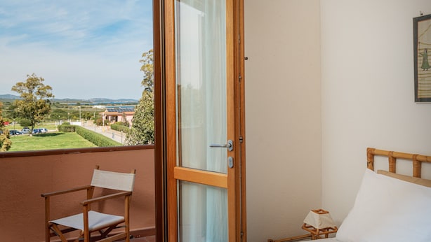 Un balcone con una sedia di legno, un letto con testiera in legno e vista su un paesaggio verde con edifici in lontananza.