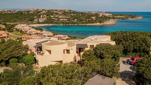 En villa vid kusten med utsikt över havet, omgiven av grönska och andra byggnader.