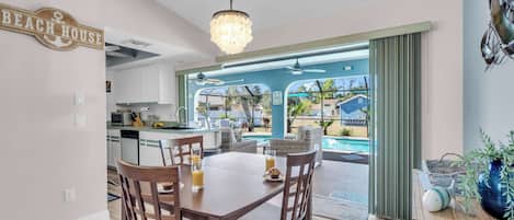 A dining area with a table and chairs, a kitchen in the background, and a view of a pool and outdoor seating area.