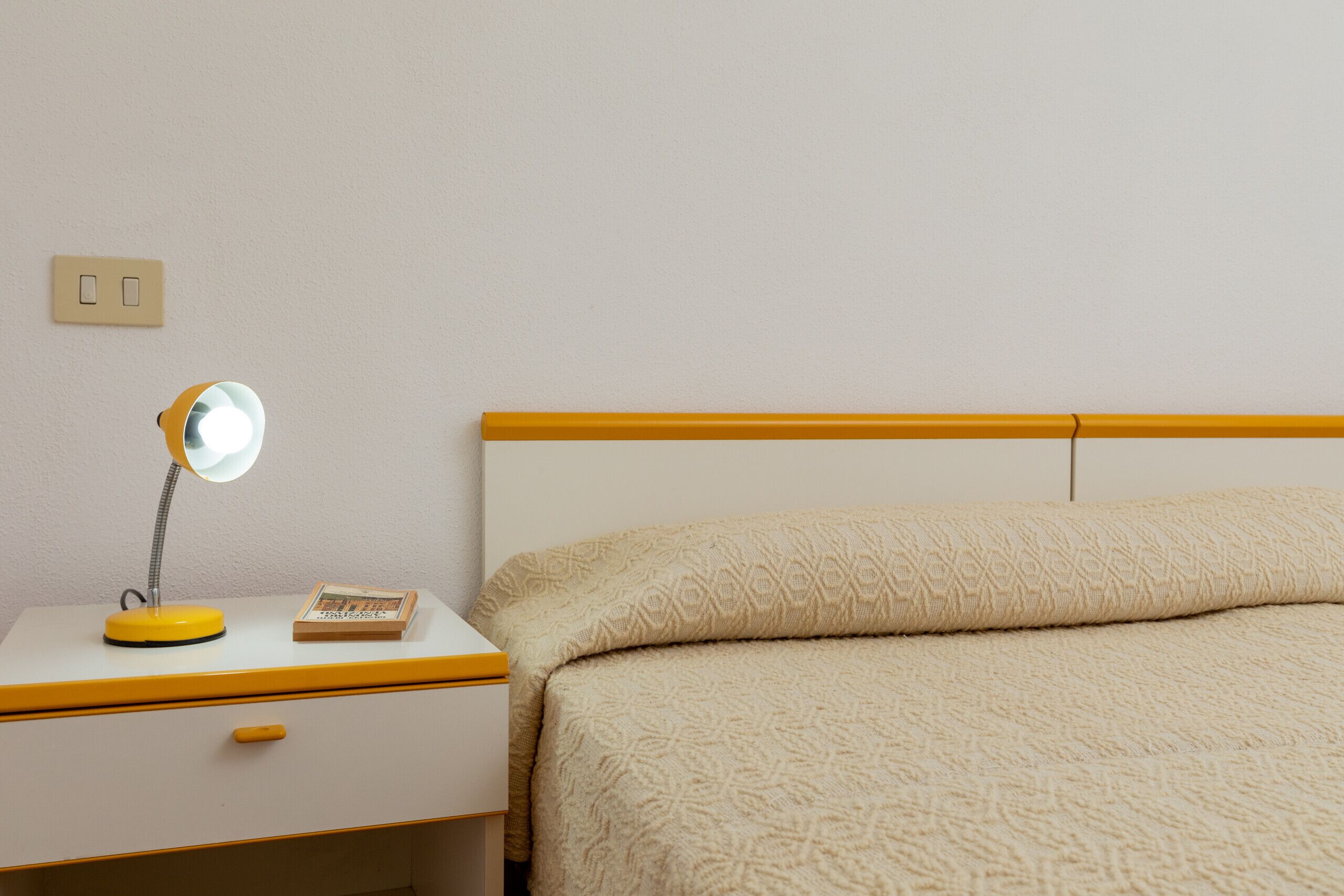 A bedroom with a bed, a nightstand, a lamp, and a wall socket.
