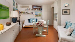 Een gezellige, goed verlichte kamer met een eettafel, stoelen, een bank en een televisie. Tegen de muren staan boekenkasten en hangt er kunst.