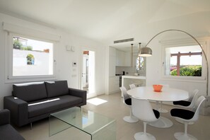 A modern living room with a glass coffee table, a grey sofa, and a white dining area with four chairs. - Villa Maddalena a Cannigione by Wonderful Italy (Arzachena)
