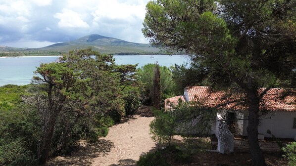 Un sentier côtier mène à un bâtiment doté d’un toit en tuiles, entouré d’une végétation luxuriante et offrant une vue sur la mer et les collines lointaines.