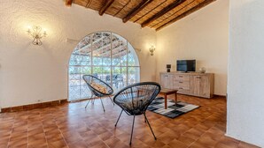 Ruime kamer met betegelde vloer, twee zwarte frame stoelen, een houten kast met televisie en een groot rondboogvenster met decoratieve ijzerwerk.
