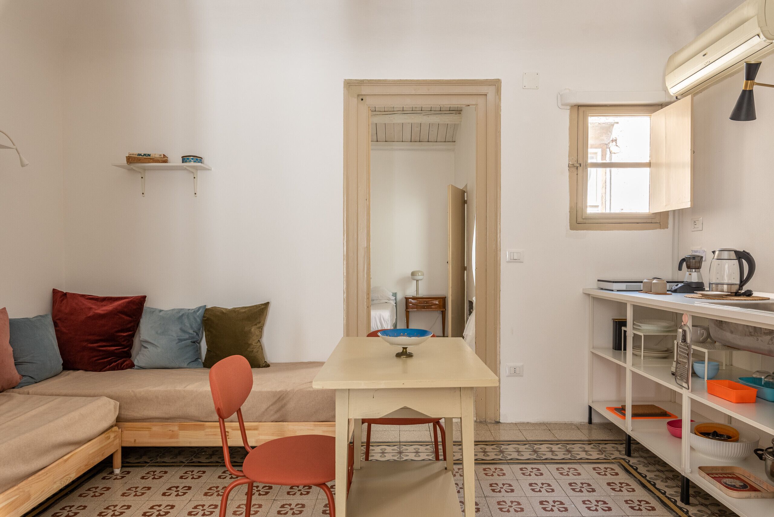 A compact living space with a sofa, a small dining table, a red chair, a kitchen area with shelves, and a patterned floor.