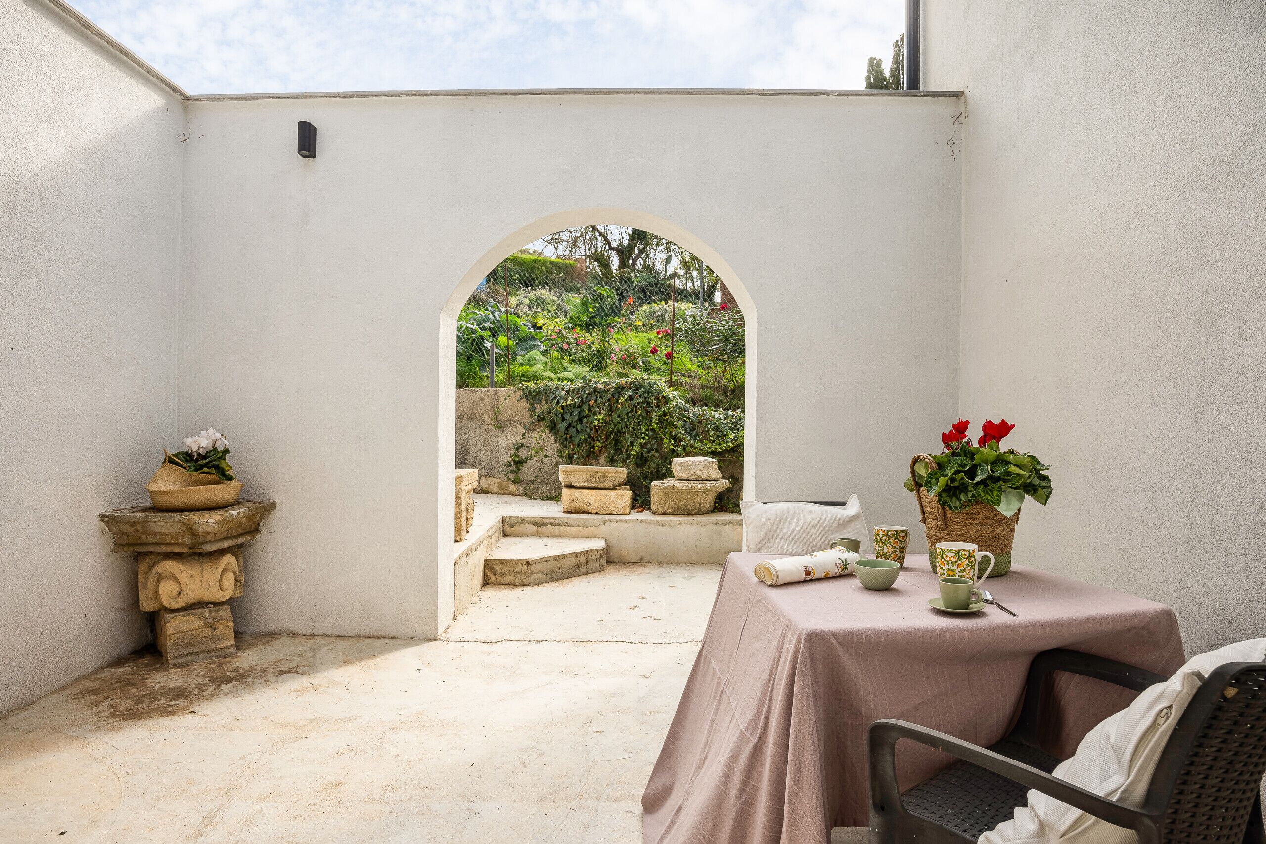A patio area with a table and chairs, a stone bench, and a view of a garden through an archway.