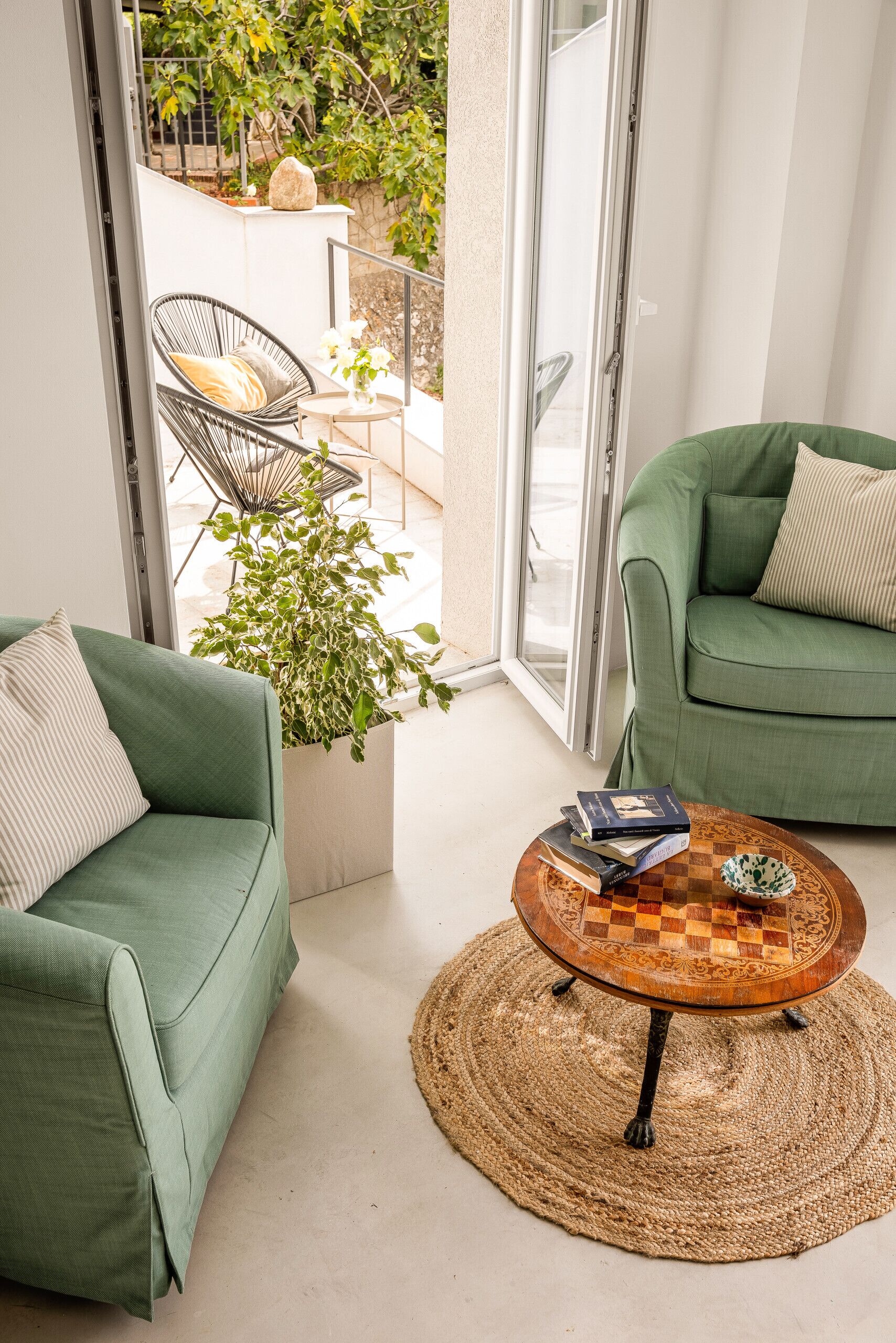 Una sala de estar moderna con un sofá verde, un sillón a juego y una mesa de centro con libros. Una planta grande en maceta se encuentra en el centro, y la habitación se abre a un balcón con vistas a la vegetación.