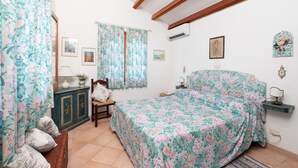 Une chambre à coucher avec une couvre-lit à motifs floraux, une tête de lit en bois, un banc, une armoire et un climatiseur fixé au mur.