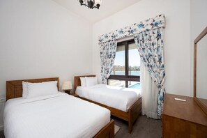 A hotel room with two single beds, a wooden headboard, a nightstand, a lamp, and a window with floral curtains. - VESTA - Gouna Residence  (El Gouna)