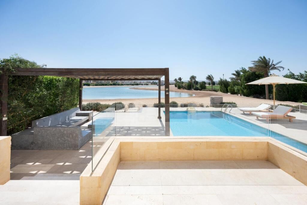 A pool area with a pergola, lounge chairs, and a view of the beach.