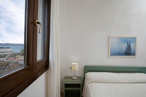 Une chambre à coucher avec un lit, une table de chevet avec une lampe, un tableau représentant un voilier et une fenêtre donnant sur la mer.