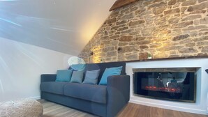 A cozy living room with a stone wall, a fireplace, and a grey sofa with blue cushions. - Cozy Studio near Quimper Train Station – Modern & Bright (Quimper)