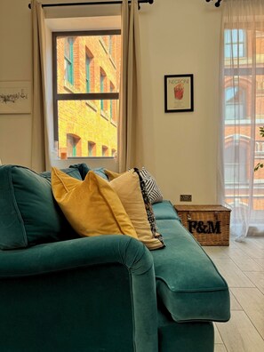 Ein blaugrünes Sofa mit Kissen, ein Korbwäscher mit der Aufschrift 'P&M' und ein Blick auf die Stadtlandschaft durch ein Fenster.