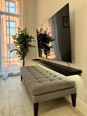 Ein modernes Wohnzimmer mit einem großen Flachbildfernseher an der Wand, einer grauen gepolsterten Sitzbank und einem Blumentopf.