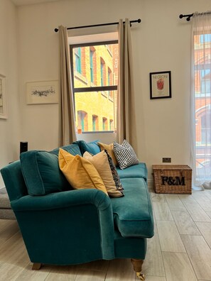 A teal sofa with cushions, a window with curtains, a framed picture on the wall, and a wicker basket on the floor. - Chambers Residence | 1BR | Prime Location (Manchester)