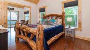 A rustic bedroom with a large bed, wooden headboard, and a view of greenery outside. - Mountain Memories-Luxury Lodge w/Views, Hot Tub (Maggie Valley)