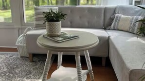 Ein modernes Wohnzimmer mit einem grauen Ecksofa, einem runden weißen Tisch mit einer Pflanze und Blick auf eine grüne Landschaft durch große Fenster.