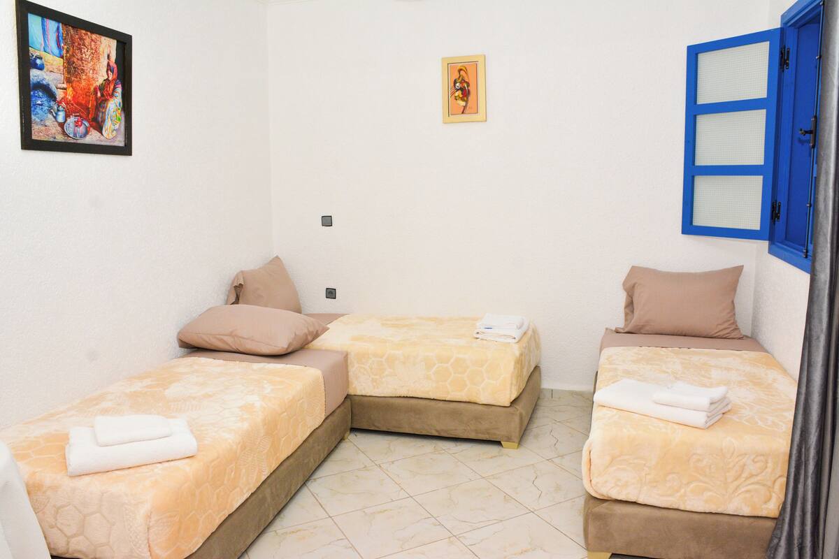 A small, clean room with two single beds, each with a pillow and a folded towel. There are two framed pictures on the wall, one of a colorful scene and another of a person.