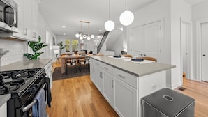 A modern kitchen with white cabinetry, a central island, and a dining area with a wooden table and chairs. - Stylish 3BR Retreat Near OSU - Sauna & Fire pit (Columbus)