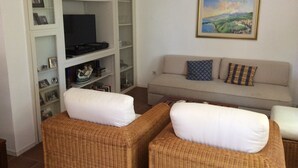 A living room with wicker furniture, a sofa with patterned cushions, a television on a shelf, and a framed landscape painting on the wall. - Apartment 'Cipolla' with Private Terrace, Private Garden and Wi-Fi (Oberkirch)