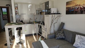 Een moderne keuken met witte kastjes, een betegelde achterwand en een eethoek met barkrukken. In de woonkamer staat een grijze bank met gekleurde kussens.