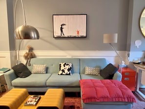 A living room with a light blue sofa, a red ottoman, and a framed artwork on the wall. - NEW! Gracious Family Townhome 18 Minutes to Manhattan (Jersey City)